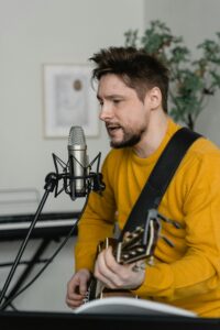 Musician in yellow sweater playing guitar and singing into a microphone indoors.