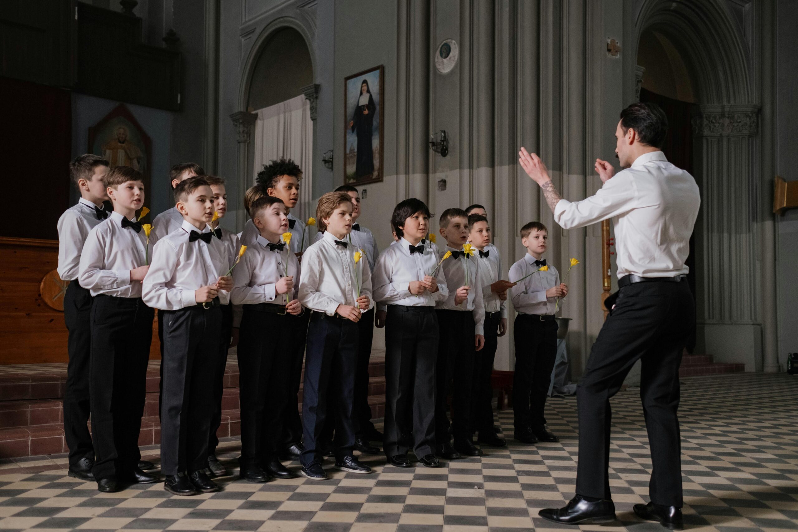 Boys choir rehearsing with a conductor indoors, holding yellow flowers.